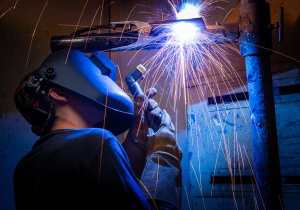 A student is wearing their welding helmet while welding. There are blue and yellow sparks flying from the scene as they work.