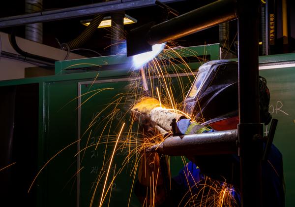A welding student is wearing full protective gear, including helmet, gloves and overalls while welding. It looks dramatic and exciting as lines of orange sparks fly from their work. 