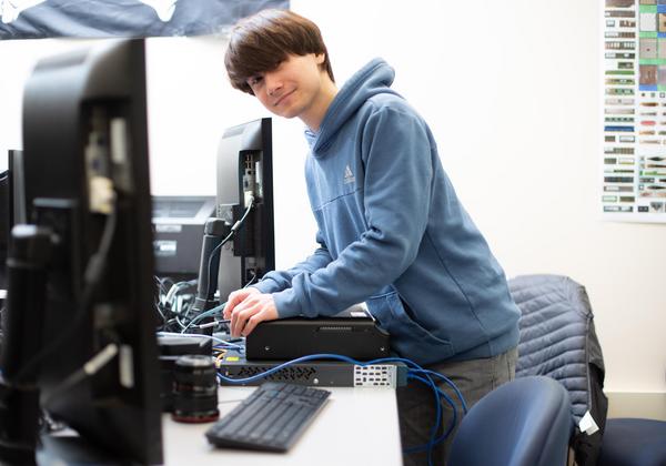 A VIU ITAS student is leaning over their desk while plugging cables into the back of a server. They are looking towards us and smiling. 