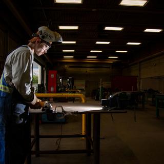 A VIU welding student who is wearing coveralls, protective head ware and gloves is preparing to start working. They are in a large, dark work space and have a spotlight on them. 