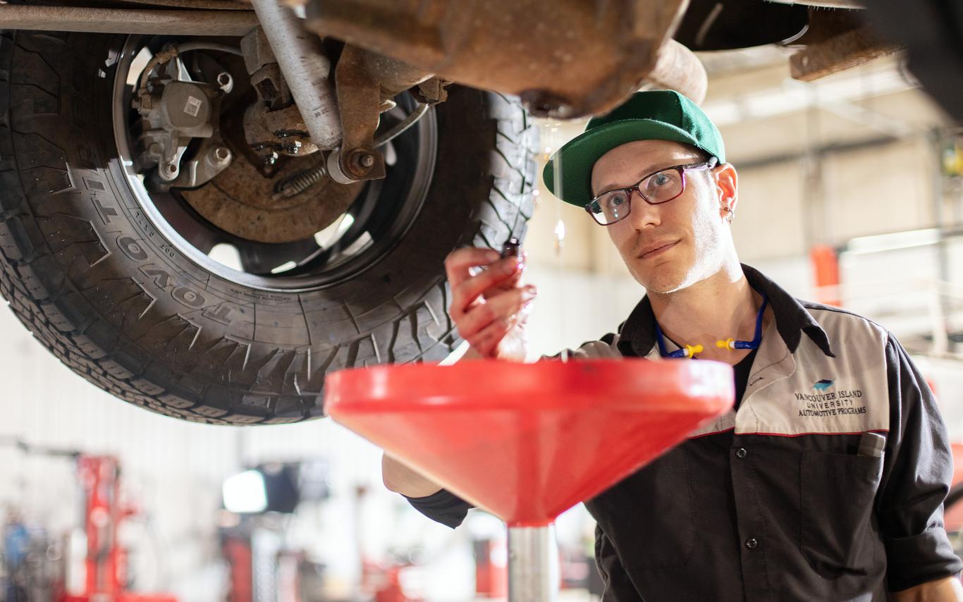 A young man in glasses working under a car on a lift. 